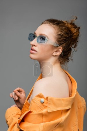 Modische junge rothaarige Frau in Sonnenbrille und orangefarbener Jeansjacke mit nackter Schulter, die wegschaut und isoliert auf grauem Hintergrund steht, trendiges Sonnenschutzkonzept, Modemodel 