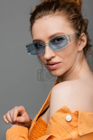 Trendiges junges sommersprossiges und rothaariges Model in blauer Sonnenbrille und orangefarbener Jeansjacke, das isoliert vor grauem Hintergrund in die Kamera blickt, trendiges Sonnenschutzkonzept 