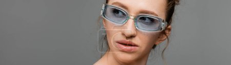 Portrait de jeune femme avec maquillage naturel et taches de rousseur portant des lunettes de soleil élégantes et regardant loin isolé sur fond gris, concept de protection solaire à la mode, bannière, modèle de mode 