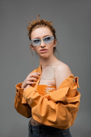 Stylish young red haired woman in sunglasses, top with sequins and orange denim jacket with naked shoulder standing isolated on grey background, trendy sun protection concept, fashion model 