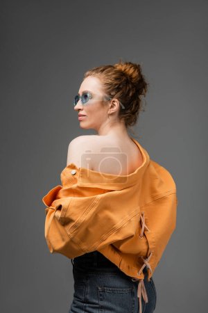 Stylische junge rothaarige Frau in Sonnenbrille und orangefarbener Jeansjacke mit nackter Schulter, die isoliert auf grauem Hintergrund steht, trendiges Sonnenschutzkonzept, Modemodel 