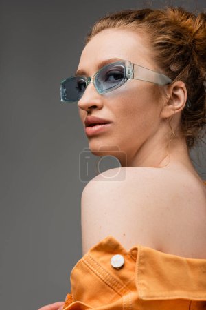 Foto de Elegante modelo pelirroja con maquillaje natural mirando a la cámara mientras posa en gafas de sol y chaqueta de mezclilla naranja con hombro desnudo aislado sobre fondo gris, concepto de protección solar de moda - Imagen libre de derechos