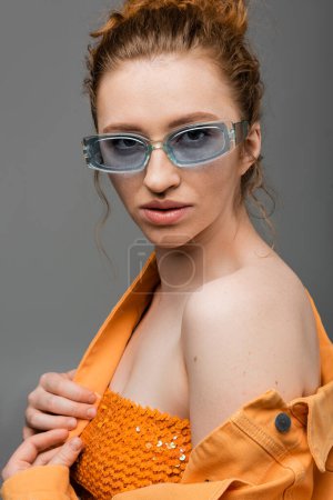 Mujer de pelo rojo joven confiada en gafas de sol posando en la parte superior con lentejuelas y chaqueta de mezclilla naranja mientras está de pie aislado sobre fondo gris, concepto de protección solar de moda, modelo de moda 