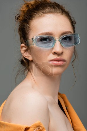 Portrait of redhead and freckled woman in sunglasses and orange denim jacket with naked shoulder looking at camera isolated on grey background, trendy sun protection concept, fashion model 