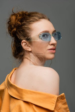 Mujer de pelo rojo moderno en gafas de sol azules y chaqueta de mezclilla naranja con hombro desnudo mirando hacia otro lado y de pie aislado sobre fondo gris, concepto de protección solar de moda 