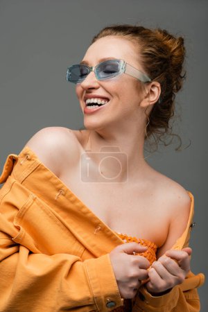 Fröhliche und stylische rothaarige Frau in blauer Sonnenbrille und orangefarbener Jeansjacke mit nackten Schultern, die isoliert auf grauem Hintergrund lachen, trendiges Sonnenschutzkonzept 