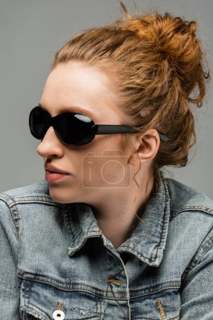 Portrait of stylish freckled and red haired woman in sunglasses and denim jacket posing and standing isolated on grey background, trendy sun protection concept, fashion model 