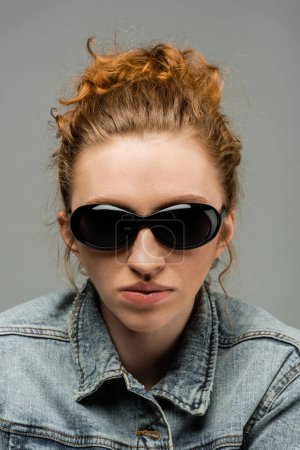 Portrait of young redhead and freckled woman with natural makeup posing in sunglasses and denim jacket while standing isolated on grey background, trendy sun protection concept, fashion model 