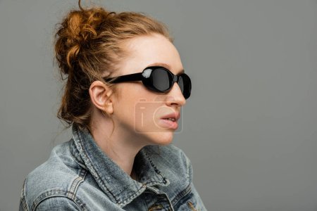 Fashionable young red haired woman with natural makeup wearing sunglasses and denim jacket while standing isolated on grey background, trendy sun protection concept, fashion model 