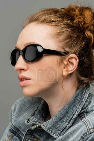 Portrait of freckled and red haired woman with natural makeup wearing stylish sunglasses and denim jacket isolated on grey background, trendy sun protection concept, fashion model 