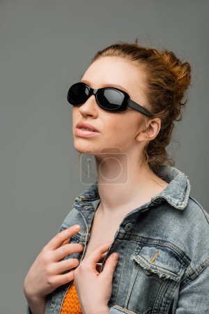 Red haired and freckled young woman in stylish sunglasses posing in denim jacket and top with sequins and standing isolated on grey background, trendy sun protection concept, fashion model 