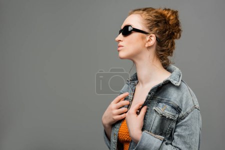 Joven modelo pelirroja con maquillaje natural posando en gafas de sol y top con lentejuelas mientras toca chaqueta de mezclilla aislada sobre fondo gris, concepto de protección solar de moda, modelo de moda 
