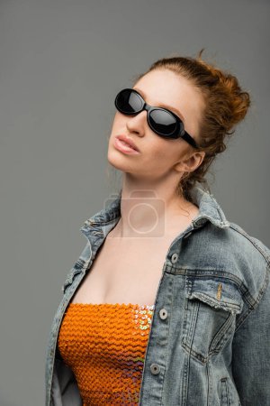 Mujer pelirroja joven confiada y elegante en gafas de sol, parte superior con lentejuelas y chaqueta de mezclilla de pie y posando aislado sobre fondo gris, concepto de protección solar de moda, modelo de moda 