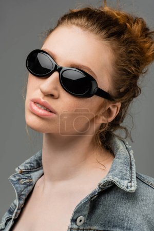 Portrait of young redhead and freckled woman in fashionable sunglasses and denim jacket standing isolated on grey background, trendy sun protection concept, fashion model 