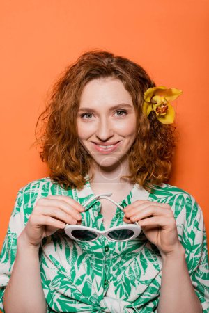 Fröhliche junge rothaarige Frau mit natürlichem Make-up und Orchideenblume im Haar, die in die Kamera schaut und Sonnenbrille auf orangefarbenem Hintergrund hält, Sommer-Freizeit- und Modekonzept, Jugendkultur