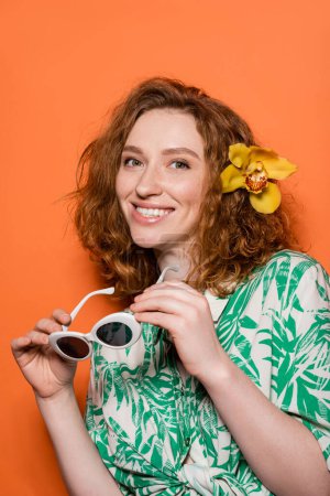 Jeune femme rousse positive avec fleur d'orchidée dans les cheveux regardant la caméra et tenant des lunettes de soleil et debout sur fond orange, concept décontracté et mode d'été, Culture de la jeunesse