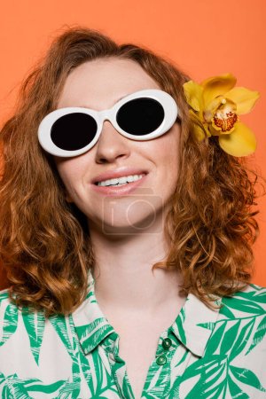 Porträt einer jungen, fröhlichen und rothaarigen Frau mit Orchideenblume im Haar, Sonnenbrille auf orangefarbenem Hintergrund stehend, Sommer-Freizeit- und Modekonzept, Jugendkultur