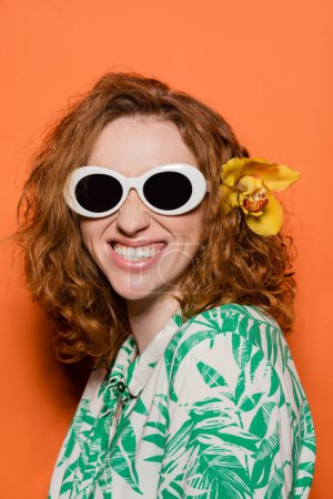 Positive Frau mit Orchideenblüte im roten Haar mit Sonnenbrille und Bluse mit floralem Muster, posierend und stehend auf orangefarbenem Hintergrund, sommerliches Freizeit- und Modekonzept, Jugendkultur