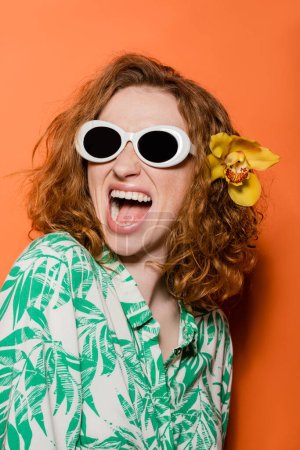 Excited redhead woman with natural makeup and orchid flower wearing sunglasses and blouse with floral print while standing on orange background, summer casual and fashion concept, Youth Culture