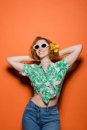 Téléchargez les photos : Joyeuse jeune femme rousse avec fleur d'orchidée dans les cheveux et les lunettes de soleil posant en chemisier avec motif floral et jeans sur fond orange, concept décontracté été et mode, Culture de la jeunesse - en image libre de droit