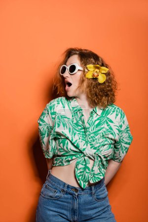 Shocked redhead woman with sunglasses and orchid flower wearing blouse with floral print and jeans while posing and standing on orange background, summer casual and fashion concept, Youth Culture