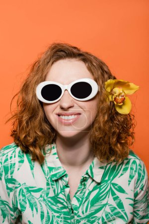 Lächelnde junge Frau mit Orchideenblüte im roten Haar mit Sonnenbrille und Bluse mit Blumenmuster, isoliert stehend auf orangefarbenem, stylischem Freizeitoutfit und sommerlichem Ambiente-Konzept, Jugendkultur