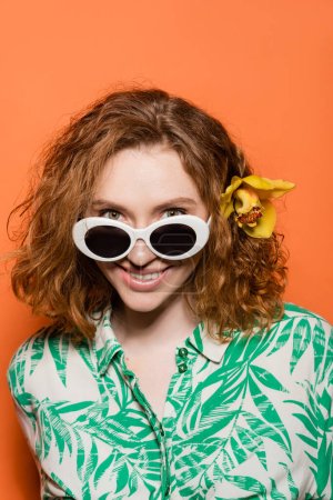 Retrato de mujer joven positiva con flor de orquídea en pelo rojo y gafas de sol mirando a la cámara mientras está de pie sobre fondo naranja, verano casual y concepto de moda, Cultura Juvenil