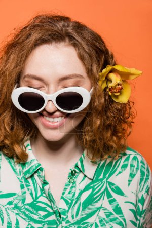 Téléchargez les photos : Joyeuse femme aux cheveux rouges avec des lunettes de soleil et une fleur d'orchidée portant une blouse à imprimé floral et se tenant isolée sur l'orange, élégante tenue décontractée et concept d'ambiance estivale, Culture de la jeunesse - en image libre de droit