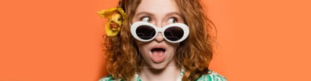 Young and shocked redhead woman with orchid flower and stylish sunglasses looking away while standing on orange background, summer casual and fashion concept, banner, Youth Culture