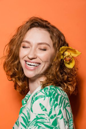 Glückliche junge Frau mit Orchideenblume im roten Haar und trendiger Bluse mit floralem Print, stehend mit geschlossenen Augen auf orangefarbenem Hintergrund, Sommer-Freizeit- und Modekonzept, Jugendkultur