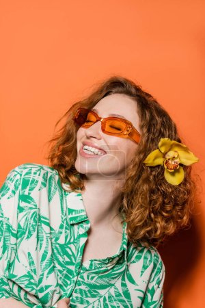 Mujer joven positiva con flor de orquídea en pelo rojo usando gafas de sol y blusa elegante mientras está de pie sobre fondo naranja, verano casual y concepto de moda, Cultura Juvenil