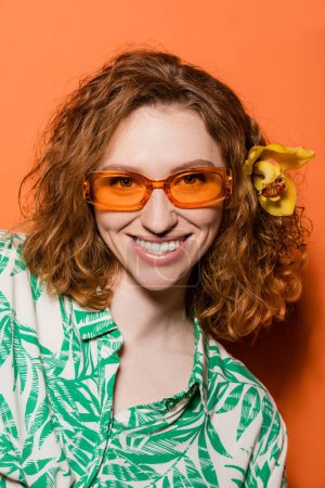 Portrait of smiling and stylish young woman with orchid flower in red hair looking at camera and posing in sunglasses on orange background, summer casual and fashion concept, Youth Culture