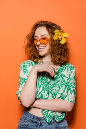 Glückliche junge rothaarige Frau mit Orchideenblume und Sonnenbrille posiert in Bluse mit floralem Print und Jeans auf orangefarbenem Hintergrund, sommerliches Freizeit- und Modekonzept, Jugendkultur