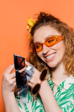 Fröhliche junge rothaarige Frau mit Orchideenblume im Haar, die eine moderne Sonnenbrille trägt und dabei isoliert auf orangefarbenem, stylischem Freizeitoutfit und sommerlichem Ambiente steht, Konzept Jugendkultur