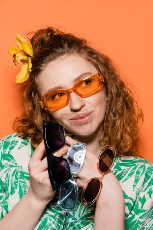 Porträt einer fröhlichen jungen Frau mit Orchideenblume im Haar, Sonnenbrille haltend und wegschauend, während sie posiert und auf orangefarbenem Hintergrund steht, Sommerleger- und Modekonzept, Jugendkultur