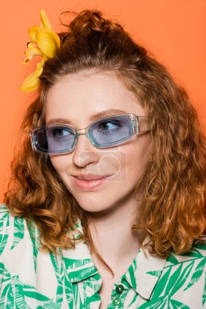 Portrait de jeune modèle rousse avec fleur d'orchidée dans les cheveux portant des lunettes de soleil bleues et chemisier à la mode avec motif floral isolé sur orange, tenue décontractée élégante et ambiance estivale concept