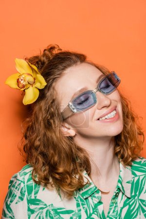Lächelnde und trendige junge Frau mit roten Haaren und Orchideenblume posiert in Sonnenbrille und Bluse mit floralem Print auf orangefarbenem Hintergrund, Sommer-Freizeit- und Modekonzept, Jugendkultur