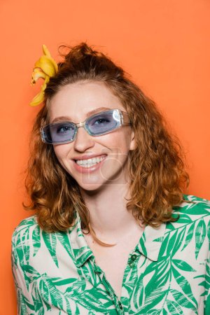Fröhliche junge rothaarige Frau mit Orchideenblüte im Haar und blauer Sonnenbrille, die isoliert auf orangefarbenem, stylischem Freizeitoutfit und sommerlichem Ambiente-Konzept steht und in die Kamera blickt