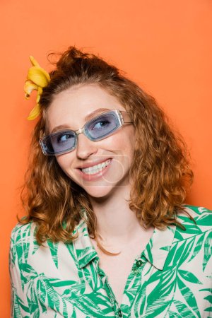 Retrato de alegre joven pelirroja con flor de orquídea en pelo y gafas de sol azules mirando hacia otro lado y de pie sobre fondo naranja, verano casual y concepto de moda 