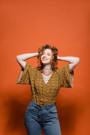 Fashionable young redhead woman in blouse with modern abstract print and jeans touching head on orange background, stylish casual outfit and summer vibes concept, Youth Culture
