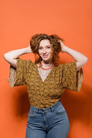 Trendige junge rothaarige Frau in moderner Bluse mit Muster und Jeans, die die Haare berühren und auf orangefarbenem Hintergrund stehen, stylisches lässiges Outfit und sommerliches Ambiente Konzept, Jugendkultur