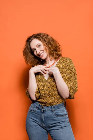 Joyful and stylish young redhead woman in blouse with abstract pattern and jeans touching necklaces and standing on orange background, stylish casual outfit and summer vibes concept, Youth Culture