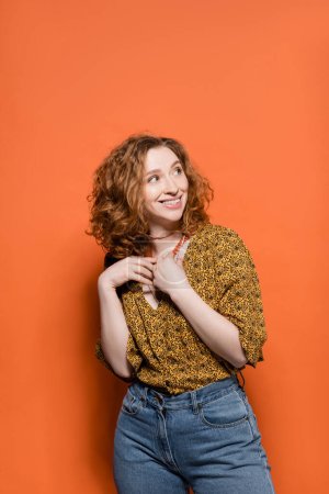 Photo for Cheerful and stylish young redhead woman in casual modern blouse and jeans touching necklaces and standing on orange background, stylish casual outfit and summer vibes concept, Youth Culture - Royalty Free Image