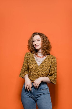 Trendiges junges rothaariges Model in gelber Bluse mit abstraktem Muster und Jeans, das in die Kamera lächelt und auf orangefarbenem Hintergrund steht, stylisches lässiges Outfit und Konzept für Sommerstimmung, Jugendkultur