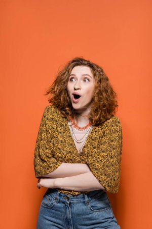 Astonished young red haired woman in yellow blouse with abstract pattern and jeans looking away while standing on orange background, stylish casual outfit and summer vibes concept, Youth Culture