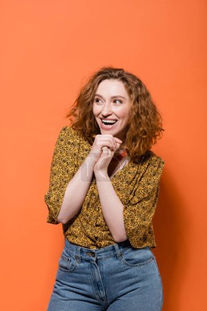 Positive young red haired woman in trendy yellow blouse with abstract pattern and jeans looking away while standing on orange background, stylish casual outfit and summer vibes concept, Youth Culture