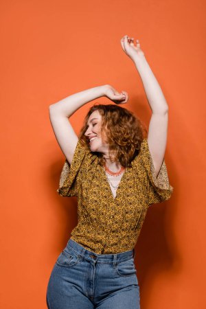 Fröhliche junge rothaarige Frau in gelber Bluse mit abstraktem Muster und Jeans, die auf orangefarbenem Hintergrund tanzt, stylisches lässiges Outfit und sommerliches Ambiente Konzept, Jugendkultur