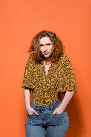 Photo for Serious young red haired model in blouse with abstract print and jeans holing hands in pockets and looking at camera on orange background, stylish casual outfit and summer vibes concept - Royalty Free Image