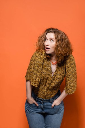 Photo for Shocked young red haired woman in yellow blouse and trendy jeans looking away while posing and standing on orange background, stylish casual outfit and summer vibes concept, Youth Culture - Royalty Free Image