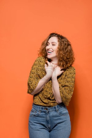 Joyful and stylish young redhead woman in yellow blouse and jeans looking away while standing on orange background, stylish casual outfit and summer vibes concept, Youth Culture